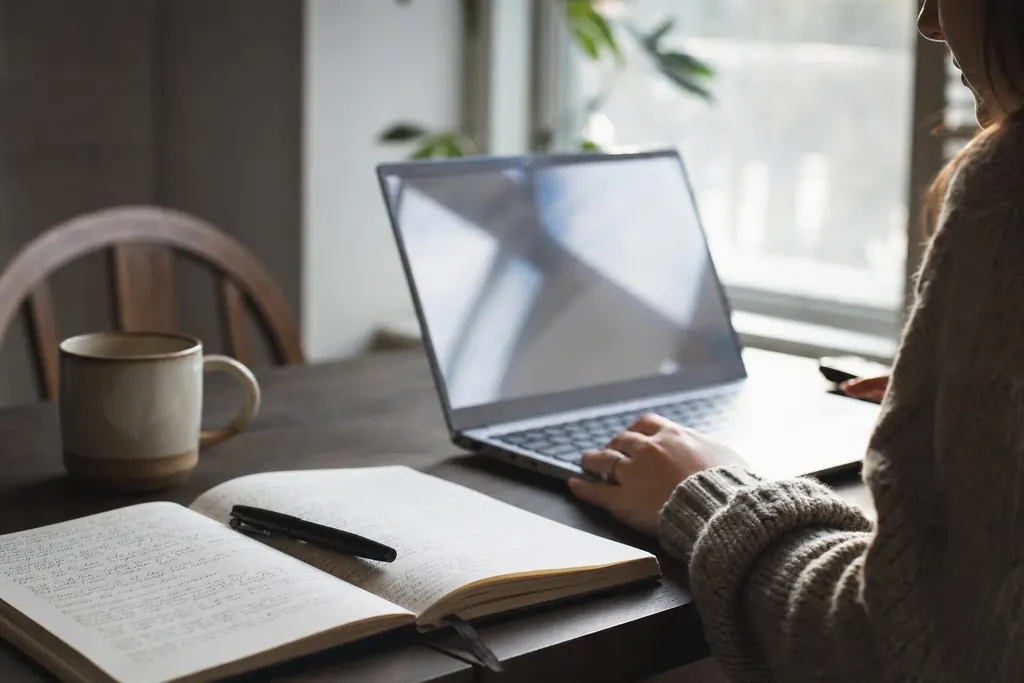 A laptop open on a table beside a handwritten notebook and a mug